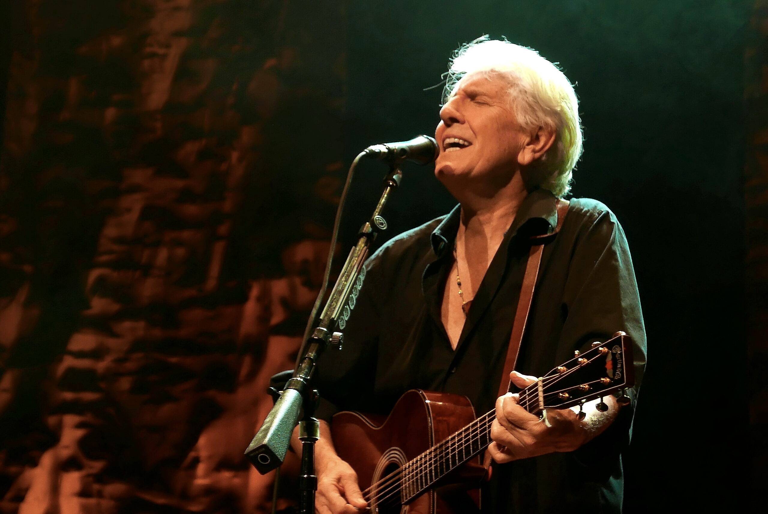 Graham Nash singing into a microphone and playing a guitar