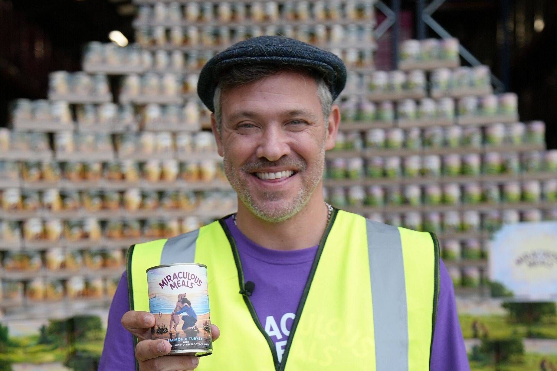 Will Young in a hi vis vest holding a tin of dog food with a wall of tins behind him