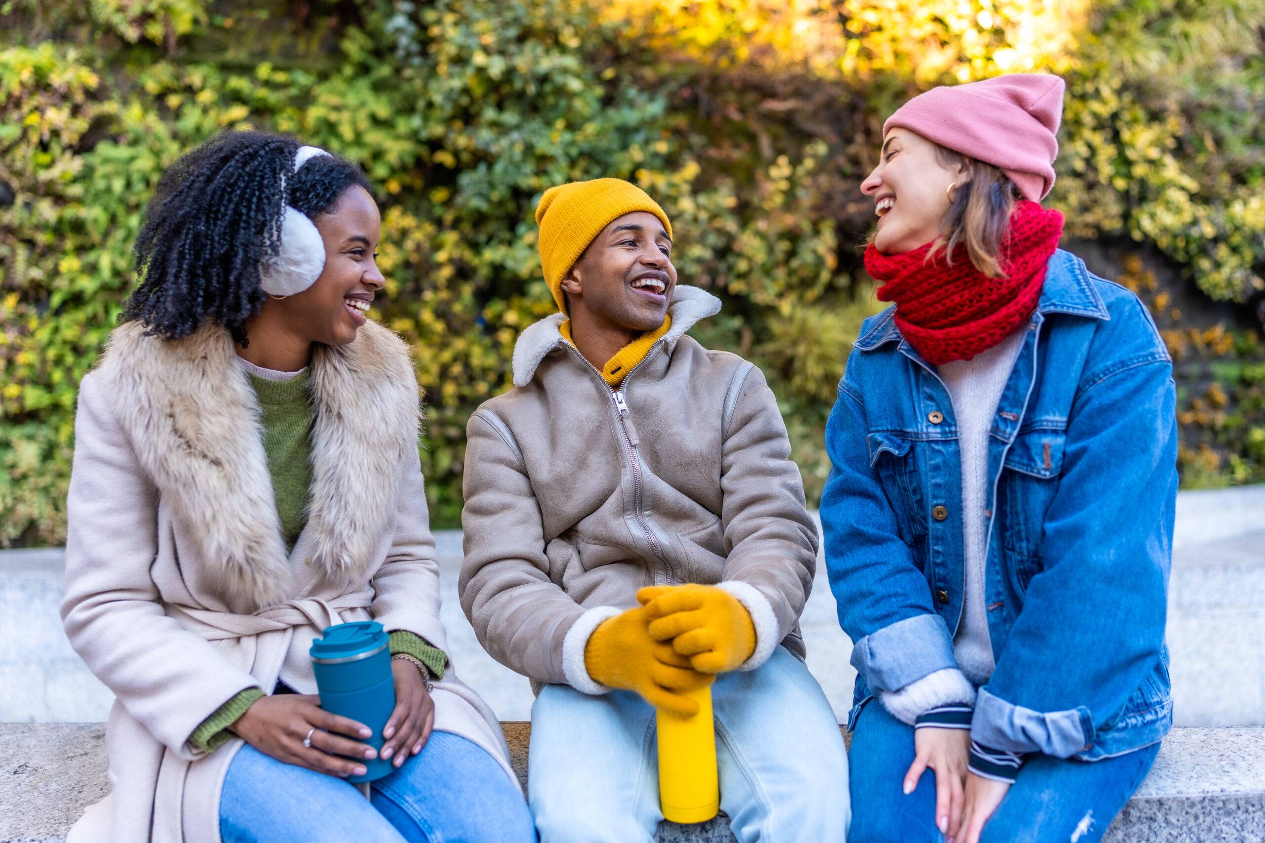 Three happy multi ethnic young friends are sitting and talking together in a city park during winter, enjoying a warm beverage