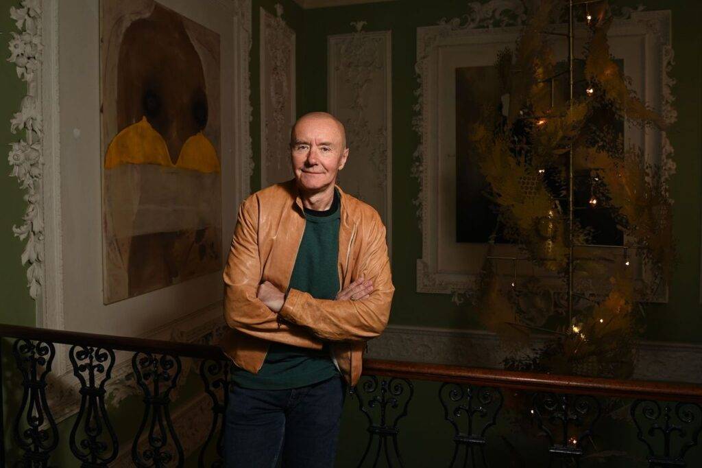 Image of Irvine Welsh leaning on a balcony railing with pictures behind him