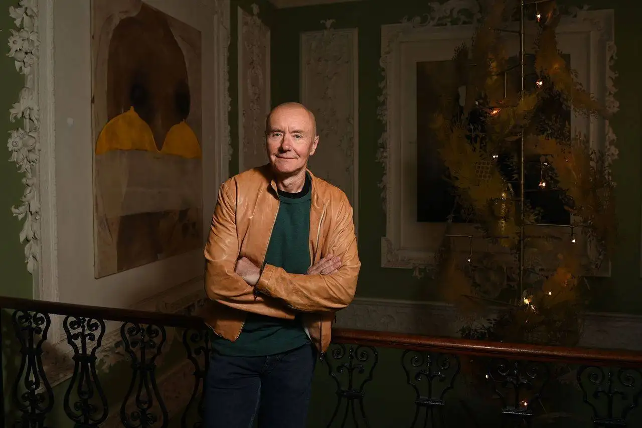 Image of Irvine Welsh leaning on a balcony railing with pictures behind him