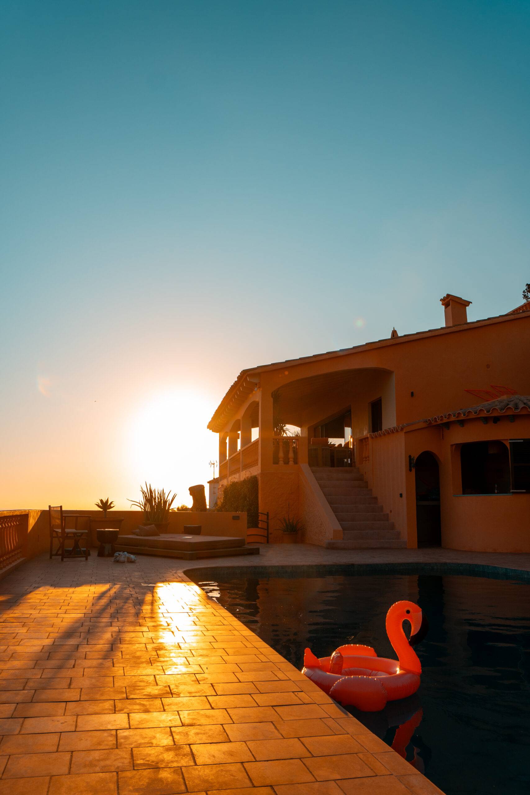 A Picture of a sun setting behind a villa and a swimming pool in the foreground of the photo