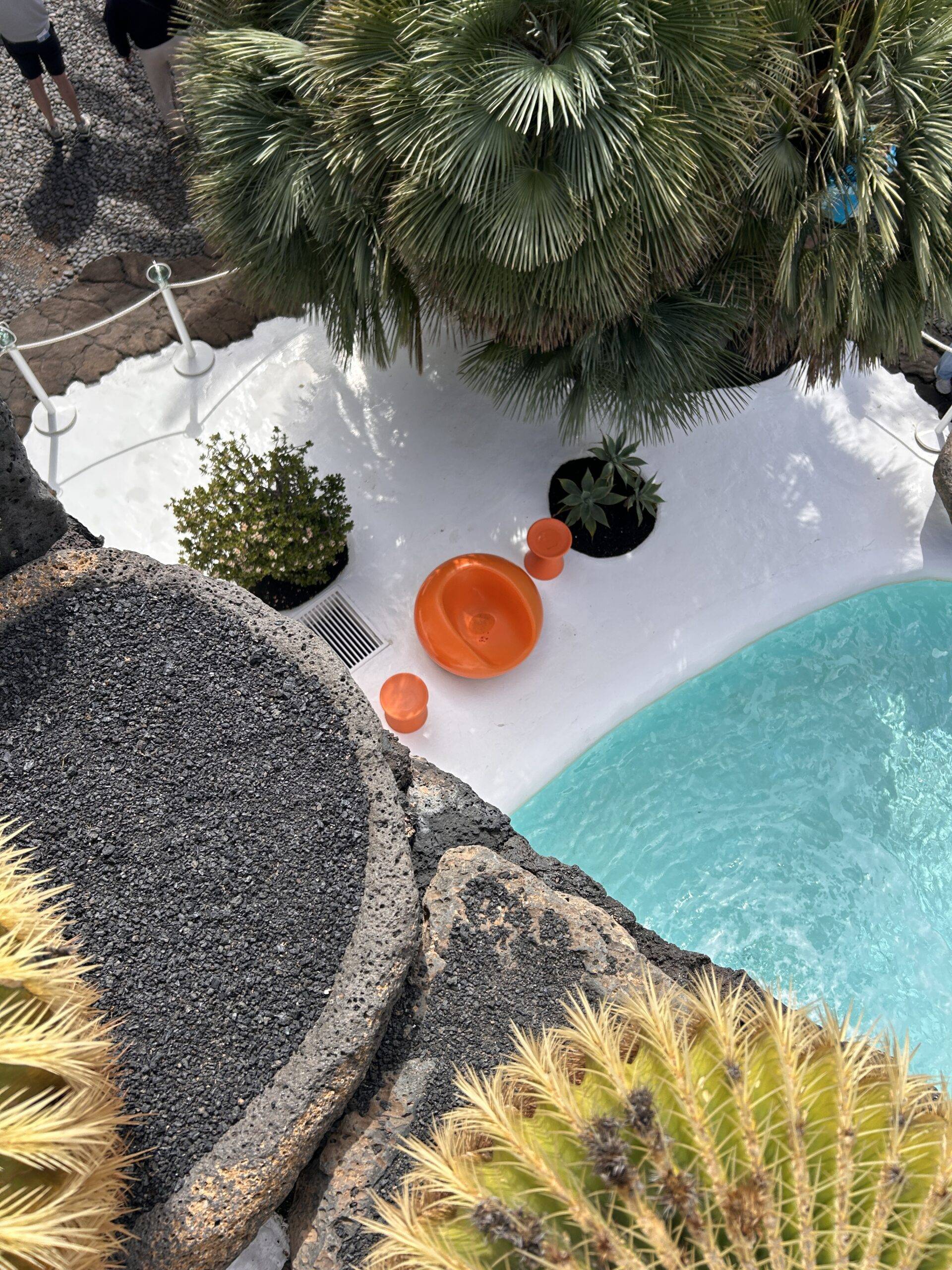 In the foreground there are black stones on top of a roof and in the background is a swimming pool with really blue water