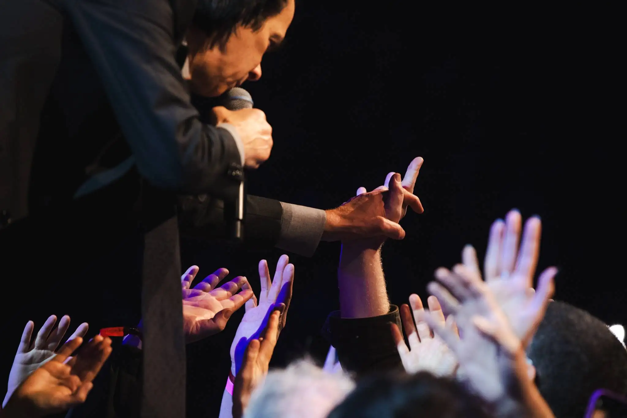 Photo of Nick Cave during a performance. He is reaching out into the crowd and holding an audience member's hand while he sings.
