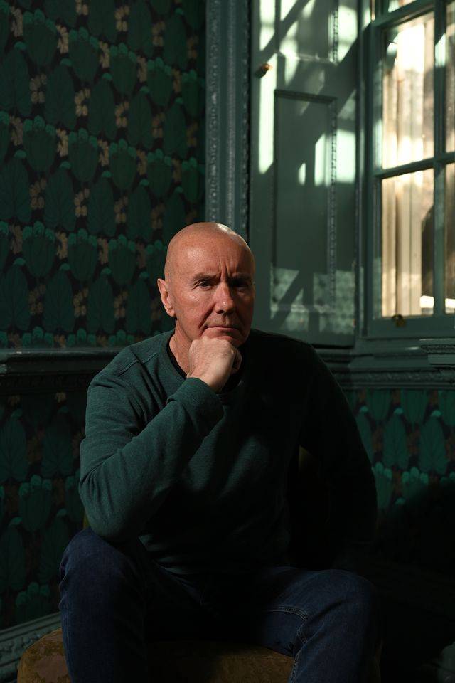 Image of Irvine Welsh sitting on a couch with light coming through the window on the right.
